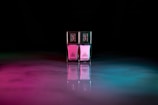 Two bottles of nail polish stand side by side, each reflecting a blend of pink and blue hues on a glossy surface. The bottles have gold caps with intricate designs, and the background is a dark, abstract space with ambient colored lighting.