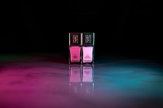 Two bottles of nail polish stand side by side, each reflecting a blend of pink and blue hues on a glossy surface. The bottles have gold caps with intricate designs, and the background is a dark, abstract space with ambient colored lighting.