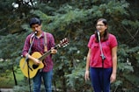 In a natural, outdoor setting, a man and a woman are performing music together. The man is playing an acoustic guitar while wearing a red checkered shirt and is singing into a microphone. The woman, wearing a pink embroidered blouse and glasses, is also singing into a microphone. They appear to be in a forested area with trees in the background.
