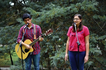 In a natural, outdoor setting, a man and a woman are performing music together. The man is playing an acoustic guitar while wearing a red checkered shirt and is singing into a microphone. The woman, wearing a pink embroidered blouse and glasses, is also singing into a microphone. They appear to be in a forested area with trees in the background.