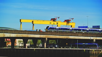 A large yellow industrial crane with the letters 'H & W' stands prominently against a clear blue sky. Below the crane, a blue train is seen traveling on an elevated railway track. Road signs and street lights are visible, adding to the urban industrial setting. Trees and building structures are partially visible under the bridge.