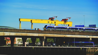 A large yellow industrial crane with the letters 'H & W' stands prominently against a clear blue sky. Below the crane, a blue train is seen traveling on an elevated railway track. Road signs and street lights are visible, adding to the urban industrial setting. Trees and building structures are partially visible under the bridge.