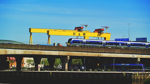 A large yellow industrial crane with the letters 'H & W' stands prominently against a clear blue sky. Below the crane, a blue train is seen traveling on an elevated railway track. Road signs and street lights are visible, adding to the urban industrial setting. Trees and building structures are partially visible under the bridge.