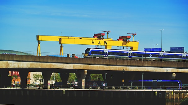 A large yellow industrial crane with the letters 'H & W' stands prominently against a clear blue sky. Below the crane, a blue train is seen traveling on an elevated railway track. Road signs and street lights are visible, adding to the urban industrial setting. Trees and building structures are partially visible under the bridge.