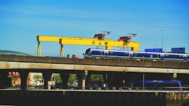 A large yellow industrial crane with the letters 'H & W' stands prominently against a clear blue sky. Below the crane, a blue train is seen traveling on an elevated railway track. Road signs and street lights are visible, adding to the urban industrial setting. Trees and building structures are partially visible under the bridge.