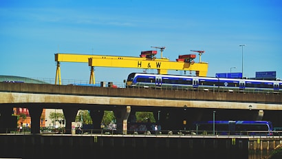 A large yellow industrial crane with the letters 'H & W' stands prominently against a clear blue sky. Below the crane, a blue train is seen traveling on an elevated railway track. Road signs and street lights are visible, adding to the urban industrial setting. Trees and building structures are partially visible under the bridge.