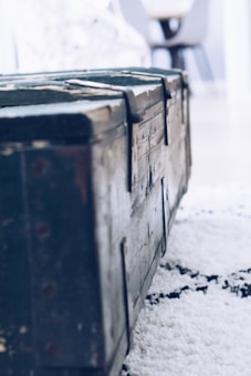 A close-up view of an old, wooden chest with metal hinges, positioned on a textured carpet. The background suggests an indoor setting with blurred furniture pieces.