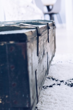 A close-up view of an old, wooden chest with metal hinges, positioned on a textured carpet. The background suggests an indoor setting with blurred furniture pieces.