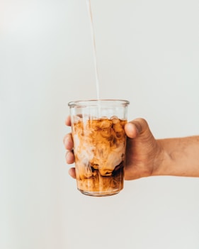 Hand hält Glas mit Eiskaffee, in das Milch eingegossen wird – stilvolle Getränkefotografie