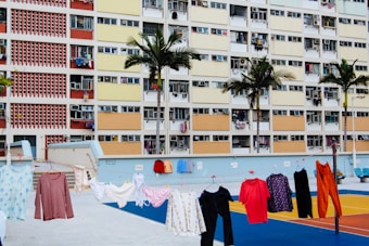 A colorful residential building with several floors and open windows, with clothes hanging out to dry on a line in front of it. The building façade displays a variety of pastel colors, including red, yellow, and orange. Palm trees are planted along the edge of the property, and there is a vibrant sports court with blue, yellow, and red sections in the foreground.