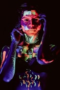 A person is covered in brightly colored fluorescent paint that glows under black light. The paint is applied in intricate patterns and designs, creating a striking and artistic effect. The background is dark, which enhances the vivid luminescence of the colors.