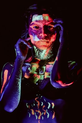 A person is covered in brightly colored fluorescent paint that glows under black light. The paint is applied in intricate patterns and designs, creating a striking and artistic effect. The background is dark, which enhances the vivid luminescence of the colors.