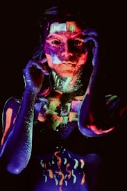 A person is covered in brightly colored fluorescent paint that glows under black light. The paint is applied in intricate patterns and designs, creating a striking and artistic effect. The background is dark, which enhances the vivid luminescence of the colors.