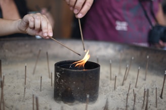 A close-up of two hands lighting incense sticks from a flame in a small, dark cylindrical container surrounded by sand. Many unlit incense sticks are standing upright in the sand, creating a serene and ritualistic atmosphere.