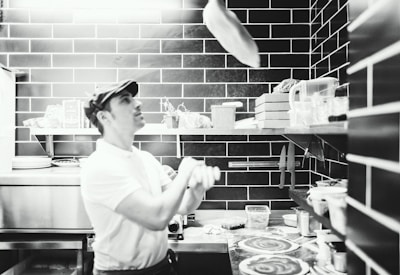 A friendly pizzaiolo tossing dough in the air inside a cozy restaurant kitchen.