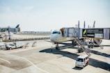 A commercial airplane is parked at an airport gate with jet bridges connected to it. Ground crew vehicles and personnel are present, including a pickup truck and a small van. The airport ramp is visible, along with distant buildings and clear skies.