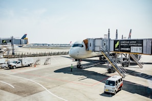 A commercial airplane is parked at an airport gate with jet bridges connected to it. Ground crew vehicles and personnel are present, including a pickup truck and a small van. The airport ramp is visible, along with distant buildings and clear skies.