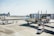 A commercial airplane is parked at an airport gate with jet bridges connected to it. Ground crew vehicles and personnel are present, including a pickup truck and a small van. The airport ramp is visible, along with distant buildings and clear skies.