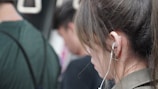 A young woman with long brown hair is seen in profile wearing earphones. She appears focused and absorbed, possibly listening to music or a podcast. Several people are in the background, suggesting a crowded environment like public transportation.