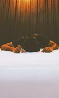 A person is resting their head on a white table with arms extended forward. The background features vertical wooden panels, creating a warm and slightly dim ambiance.