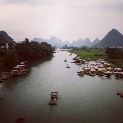 A serene river scene with multiple bamboo rafts carrying people floating gently on the water. Lush green vegetation lines the riverbanks, and in the background, a range of karst mountains is visible under a cloudy sky. Numerous colorful umbrellas adorn the resting rafts along the shore, adding vibrancy to the tranquil landscape.