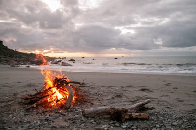 A small bonfire made of wooden logs burns brightly on a sandy beach, with waves gently lapping at the shore. The sky is overcast with thick clouds, casting a tranquil, muted light over the landscape.
