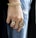 A close-up of a hand adorned with multiple pieces of jewelry including a chunky gold bracelet and two large rings. The hand rests against a pair of distressed blue jeans, creating a contrast between the luxurious gold accessories and casual denim fabric.