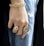 A close-up of a hand adorned with multiple pieces of jewelry including a chunky gold bracelet and two large rings. The hand rests against a pair of distressed blue jeans, creating a contrast between the luxurious gold accessories and casual denim fabric.