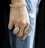 A close-up of a hand adorned with multiple pieces of jewelry including a chunky gold bracelet and two large rings. The hand rests against a pair of distressed blue jeans, creating a contrast between the luxurious gold accessories and casual denim fabric.