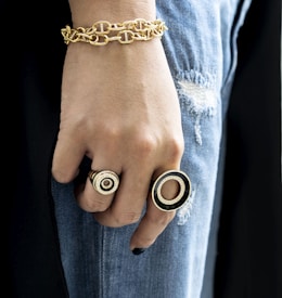 A close-up of a hand adorned with multiple pieces of jewelry including a chunky gold bracelet and two large rings. The hand rests against a pair of distressed blue jeans, creating a contrast between the luxurious gold accessories and casual denim fabric.