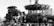 A large mechanical elephant with passengers is walking beside a carousel with intricate details. A crowd of people is gathered around these attractions, suggesting a lively public event or fair. The structures appear to be made with industrial elements and have a steampunk aesthetic.