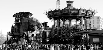 A large mechanical elephant with passengers is walking beside a carousel with intricate details. A crowd of people is gathered around these attractions, suggesting a lively public event or fair. The structures appear to be made with industrial elements and have a steampunk aesthetic.