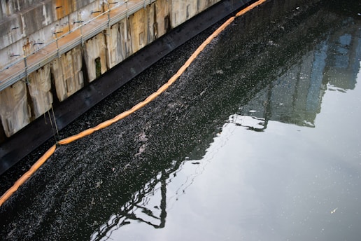 A concrete structure runs along the left side with visible metal railings. Below the structure, a layer of dark, polluted water is partially contained by a floating boom. The surface of the water reflects parts of the surroundings, suggesting an industrial environment.