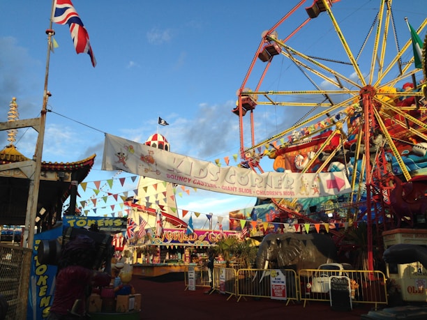 A vibrant amusement park scene featuring a large Ferris wheel with colorful decorations. Flags and multicolored bunting are strung across the area, adding to the festive atmosphere. Various rides and attractions are visible, including a carousel and a children's area marked 'KIDS CORNER' with signage for bouncy castles and slides. An elephant ride is also present, along with a 'World of Funfair' sign. The sky is clear, enhancing the colors and cheerful mood.