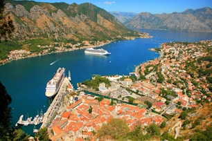A scenic coastal area with rugged mountains surrounding a winding bay. Two large cruise ships are anchored in the vibrant blue water, near a town with red-roofed buildings. Numerous smaller boats are docked along the marina. The landscape is lush with greenery scattered among the urban areas.