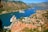 A scenic coastal area with rugged mountains surrounding a winding bay. Two large cruise ships are anchored in the vibrant blue water, near a town with red-roofed buildings. Numerous smaller boats are docked along the marina. The landscape is lush with greenery scattered among the urban areas.