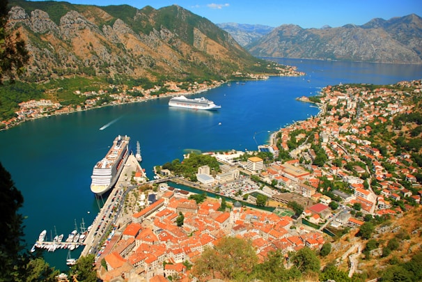 A scenic coastal area with rugged mountains surrounding a winding bay. Two large cruise ships are anchored in the vibrant blue water, near a town with red-roofed buildings. Numerous smaller boats are docked along the marina. The landscape is lush with greenery scattered among the urban areas.