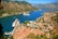 A scenic coastal area with rugged mountains surrounding a winding bay. Two large cruise ships are anchored in the vibrant blue water, near a town with red-roofed buildings. Numerous smaller boats are docked along the marina. The landscape is lush with greenery scattered among the urban areas.