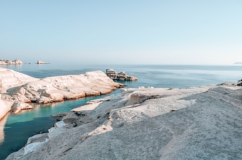 A serene coastal landscape featuring smooth white rock formations surrounded by clear blue water. The horizon line divides the calm sea from the sky, creating a tranquil and expansive view. The area appears untouched, evoking a sense of calm and solitude.