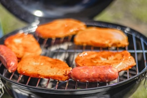 Several pieces of meat, including sausages and steak, are being grilled on an open barbecue. The meat is coated with a reddish marinade, sizzling over a flame. The background is slightly blurred, focusing attention on the food.
