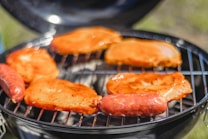 Several pieces of meat, including sausages and steak, are being grilled on an open barbecue. The meat is coated with a reddish marinade, sizzling over a flame. The background is slightly blurred, focusing attention on the food.