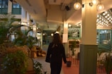 A person walks through a well-lit, indoor space featuring numerous plants and green accents. The area is adorned with hanging lamps and light fixtures, and there are decorative elements such as baskets placed along the walls. The ambiance suggests a cozy, inviting environment with a blend of natural and modern design elements.