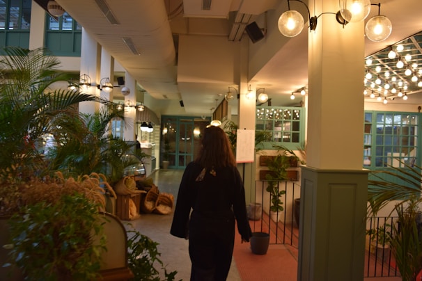 A person walks through a well-lit, indoor space featuring numerous plants and green accents. The area is adorned with hanging lamps and light fixtures, and there are decorative elements such as baskets placed along the walls. The ambiance suggests a cozy, inviting environment with a blend of natural and modern design elements.