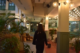 A person walks through a well-lit, indoor space featuring numerous plants and green accents. The area is adorned with hanging lamps and light fixtures, and there are decorative elements such as baskets placed along the walls. The ambiance suggests a cozy, inviting environment with a blend of natural and modern design elements.