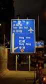A blue highway sign is prominently displayed at night, directing travelers to various locations such as Yau Ma Tei, Tsim Sha Tsui, Hung Hom, and Hong Kong. The sign includes an airplane symbol, indicating directions to an airport. It is illuminated by nearby streetlights and is positioned on a sidewalk beside a fenced construction area.