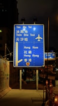 A blue highway sign is prominently displayed at night, directing travelers to various locations such as Yau Ma Tei, Tsim Sha Tsui, Hung Hom, and Hong Kong. The sign includes an airplane symbol, indicating directions to an airport. It is illuminated by nearby streetlights and is positioned on a sidewalk beside a fenced construction area.