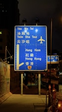A blue highway sign is prominently displayed at night, directing travelers to various locations such as Yau Ma Tei, Tsim Sha Tsui, Hung Hom, and Hong Kong. The sign includes an airplane symbol, indicating directions to an airport. It is illuminated by nearby streetlights and is positioned on a sidewalk beside a fenced construction area.