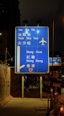 A blue highway sign is prominently displayed at night, directing travelers to various locations such as Yau Ma Tei, Tsim Sha Tsui, Hung Hom, and Hong Kong. The sign includes an airplane symbol, indicating directions to an airport. It is illuminated by nearby streetlights and is positioned on a sidewalk beside a fenced construction area.