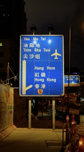 A blue highway sign is prominently displayed at night, directing travelers to various locations such as Yau Ma Tei, Tsim Sha Tsui, Hung Hom, and Hong Kong. The sign includes an airplane symbol, indicating directions to an airport. It is illuminated by nearby streetlights and is positioned on a sidewalk beside a fenced construction area.