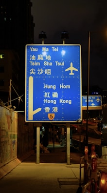 A blue highway sign is prominently displayed at night, directing travelers to various locations such as Yau Ma Tei, Tsim Sha Tsui, Hung Hom, and Hong Kong. The sign includes an airplane symbol, indicating directions to an airport. It is illuminated by nearby streetlights and is positioned on a sidewalk beside a fenced construction area.